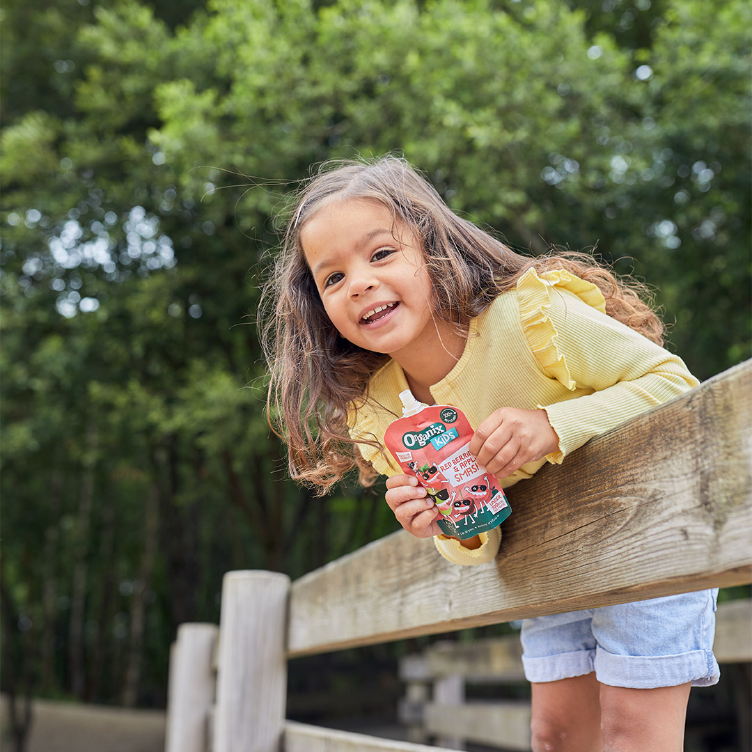 little girl with brown hair in a yellow cardigan is leaning over a wooden fence holding a pouch standing in front of a green tree