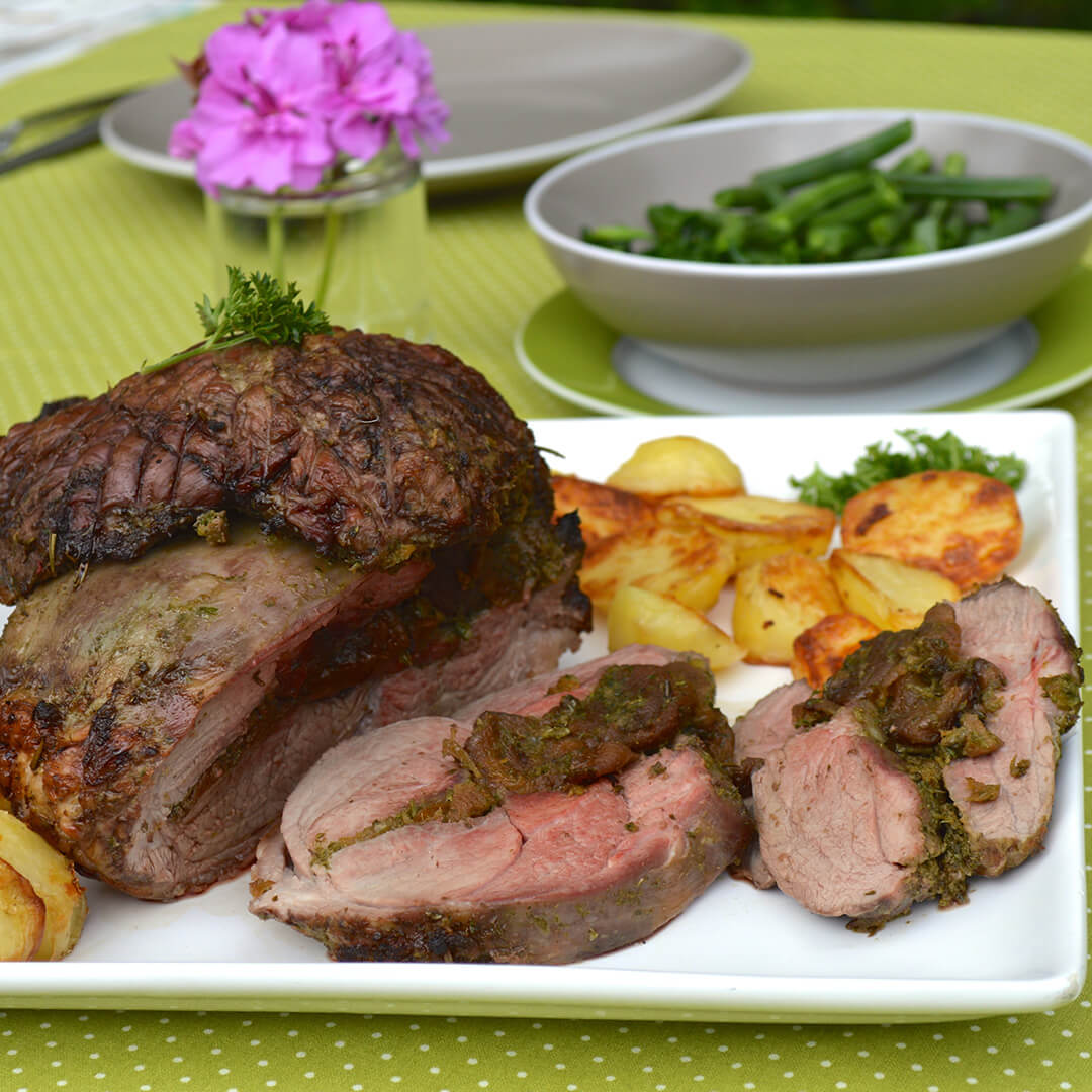 Apricot stuffed lamb on a serving dish with roast potatoes and a bowl of green beans next to it