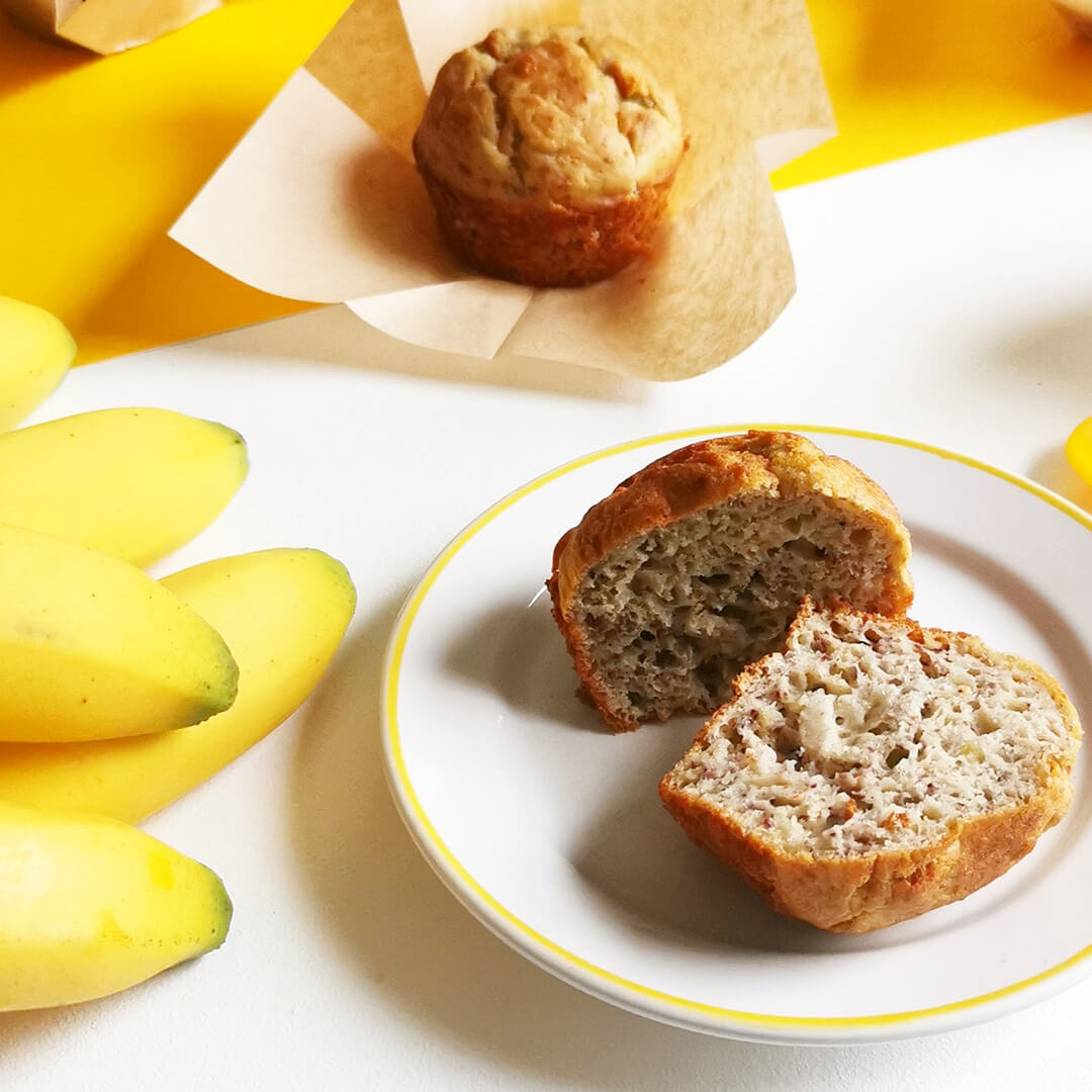 Baby banana muffins, one cut in half on a plate, the other on a paper cake case next to a bunch of bananas