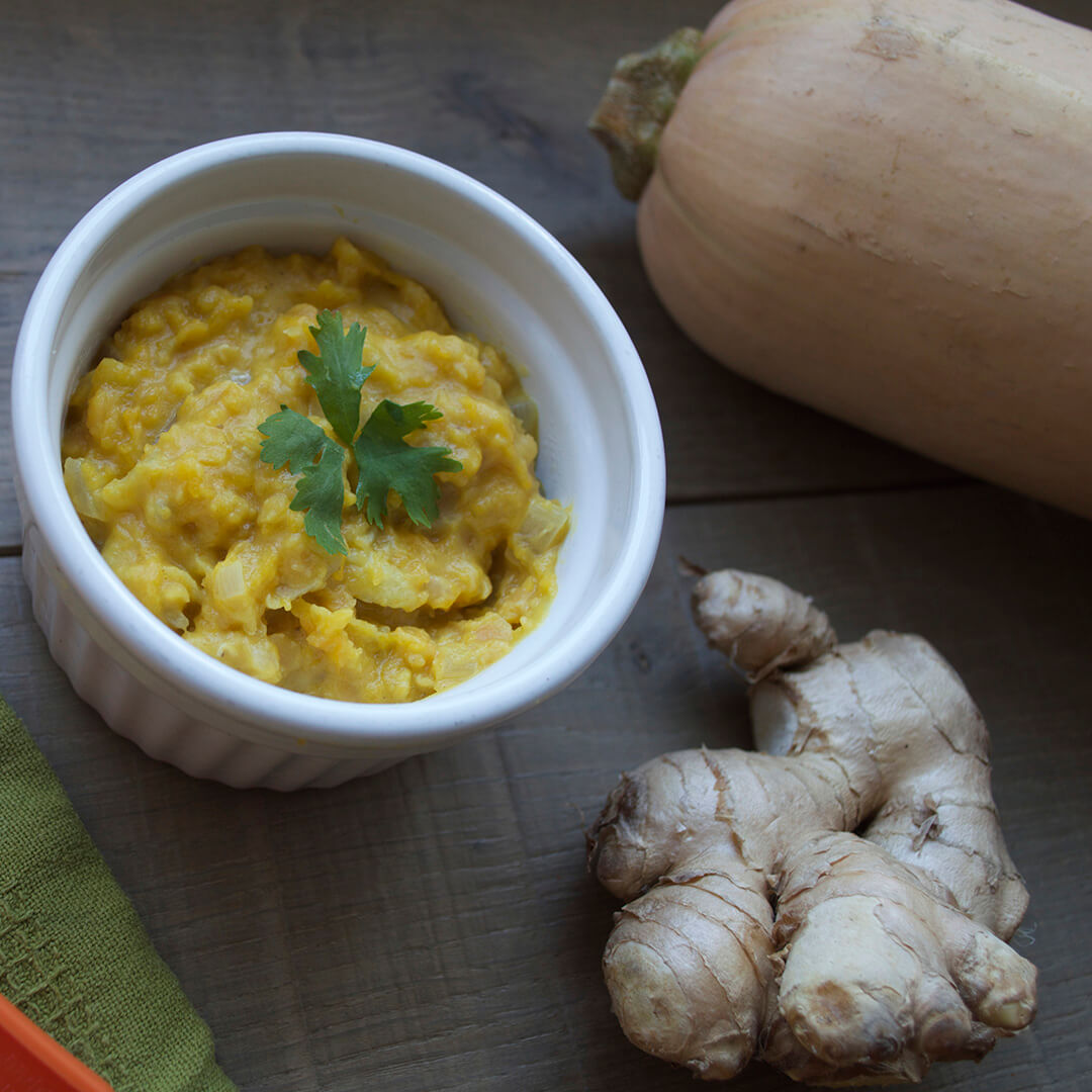 A bow of butternut squash & ginger puree next to a ginger root and butternut squash