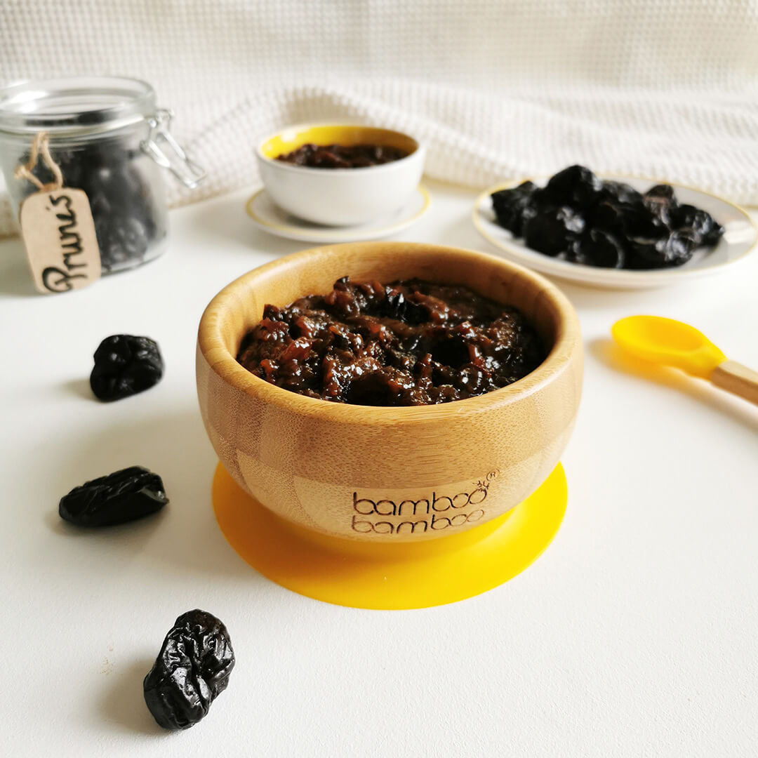 A small bowl of prune puree with some prunes around it and next to a jar of prunes, a small plate of prunes