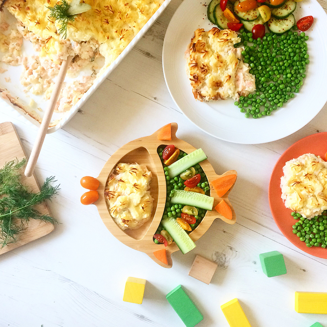 A serving of Salmon & Leek Fish Pie for Baby with cucumber sticks and veggies, two other serving with peas and a casserole dish with the fish pie