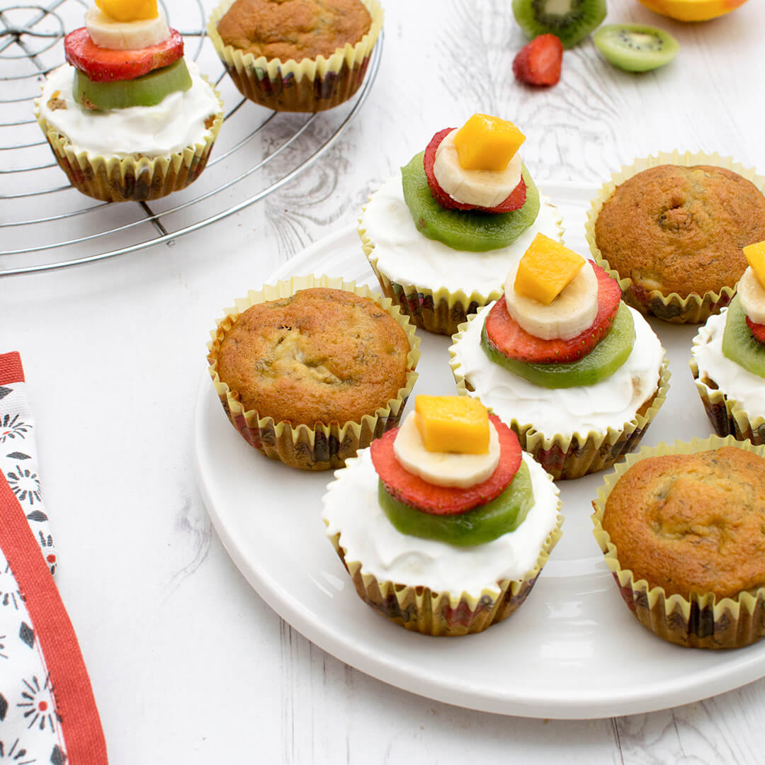 A plate of Gummy banana cupcakes, some un-topped/undecorated and some decorated with Greek yoghurt and fruit segments/slices