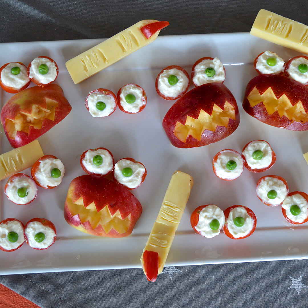 A platter of Halloween snacks: cherry tomatoes topped with cream cheese and peas to make eyes, cheese sticks with red pepper to make fingers with red nails and apple slices cut to look like mouths