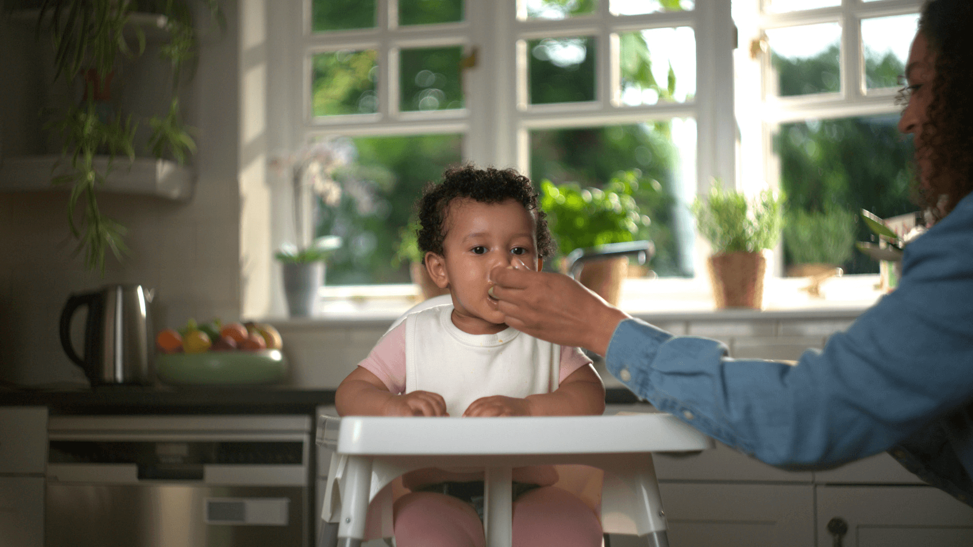 Baby in a kitchen, on a highchair, eating food being fed by mother