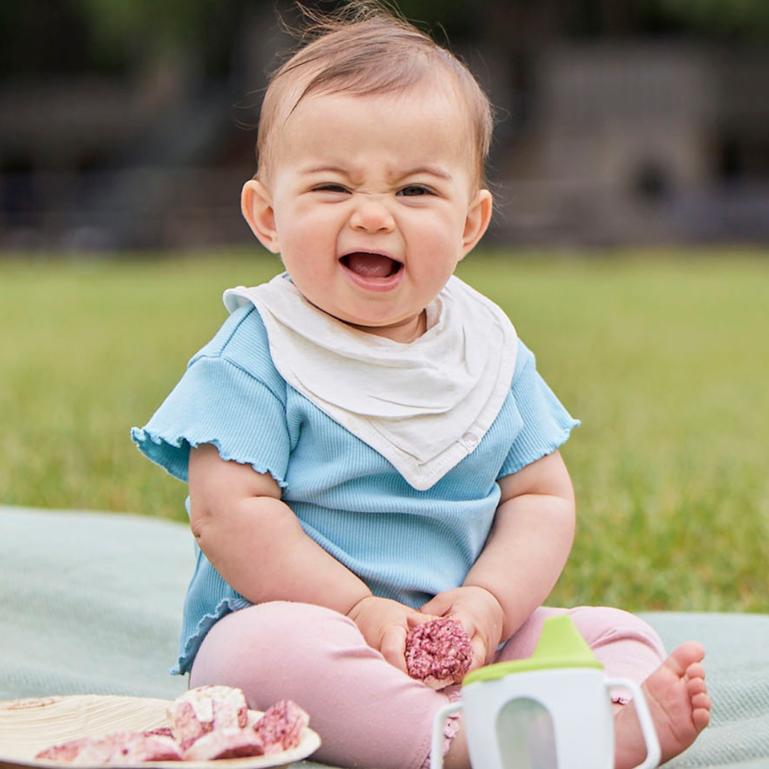 Baby sitting outdoors on a picnic blanket eating Organix Rice Cake Clouds