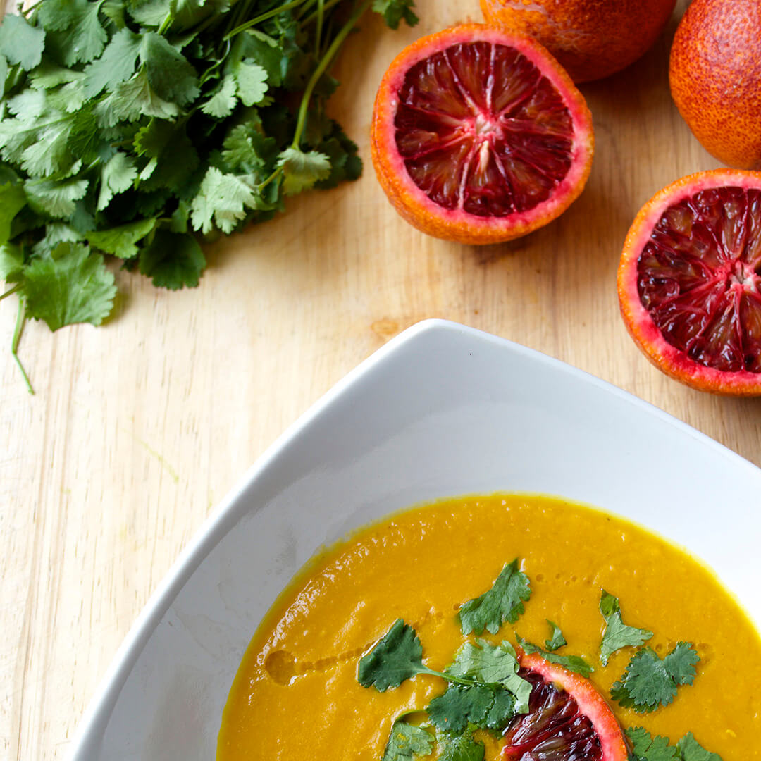 A bowl of Blood Orange, Red Lentil & Carrot Soup next to halved blood oranges and a bunch of coriander