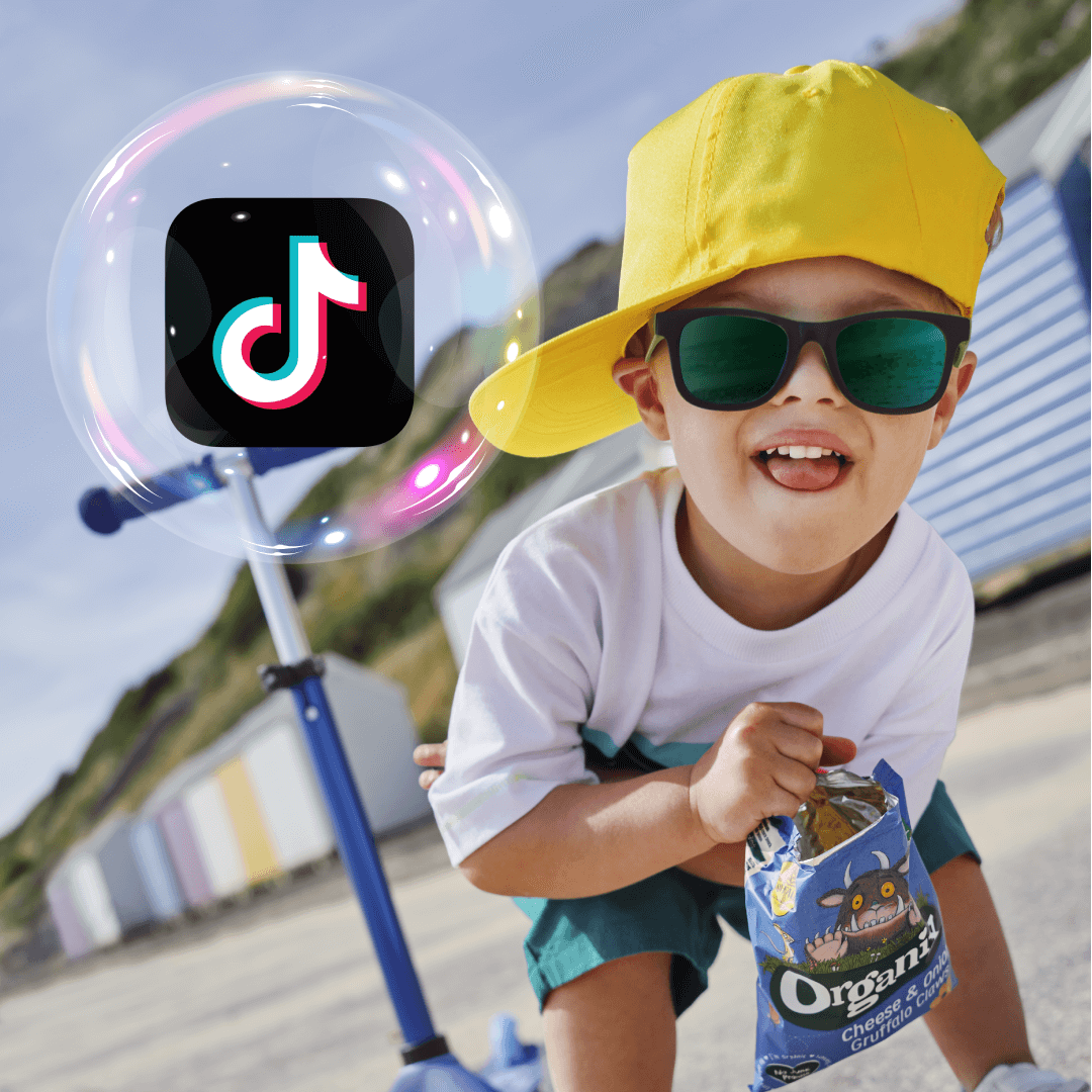 A child at the beach wearing a bright yellow cap sideways and sunglasses, with a cool expression and holding a bag of Organix Gruffalo claws (puffs). Also the TikTok logo inside an Organix Lil' Ballers bubble. And standing next to a child scooter.