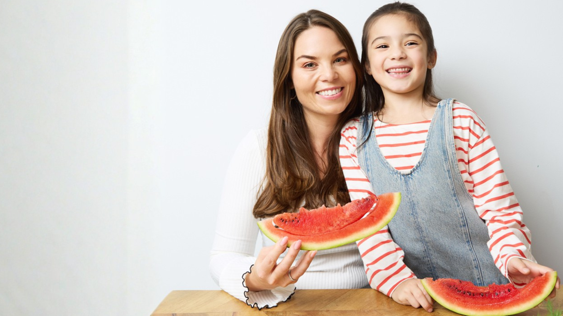 Image of a young mother and her child, holding watermelon slices. Sitting at a wooden table. Both people are smiling and looking directly at the camera. 