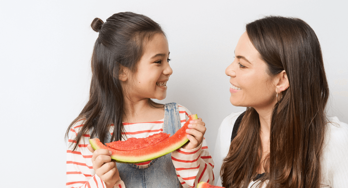 6 year old girl and mum with long dark brown hair are smiling at each other whilst the daughter holds a half eaten watermelon.