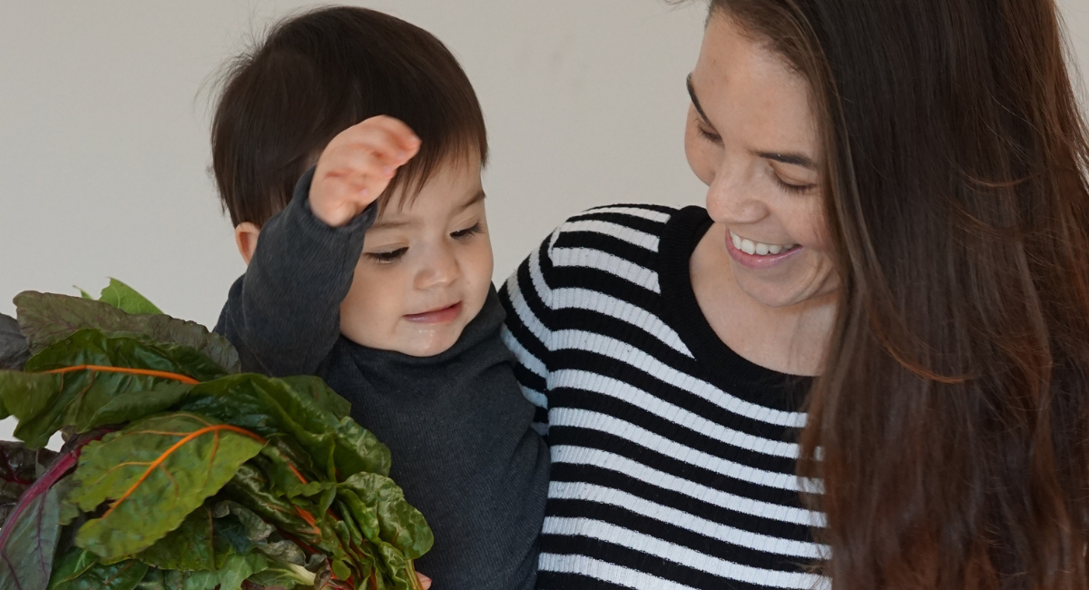 Mum in a stripy black and white top is holding baby daughter with dark brown hair and carrying some beetroot.