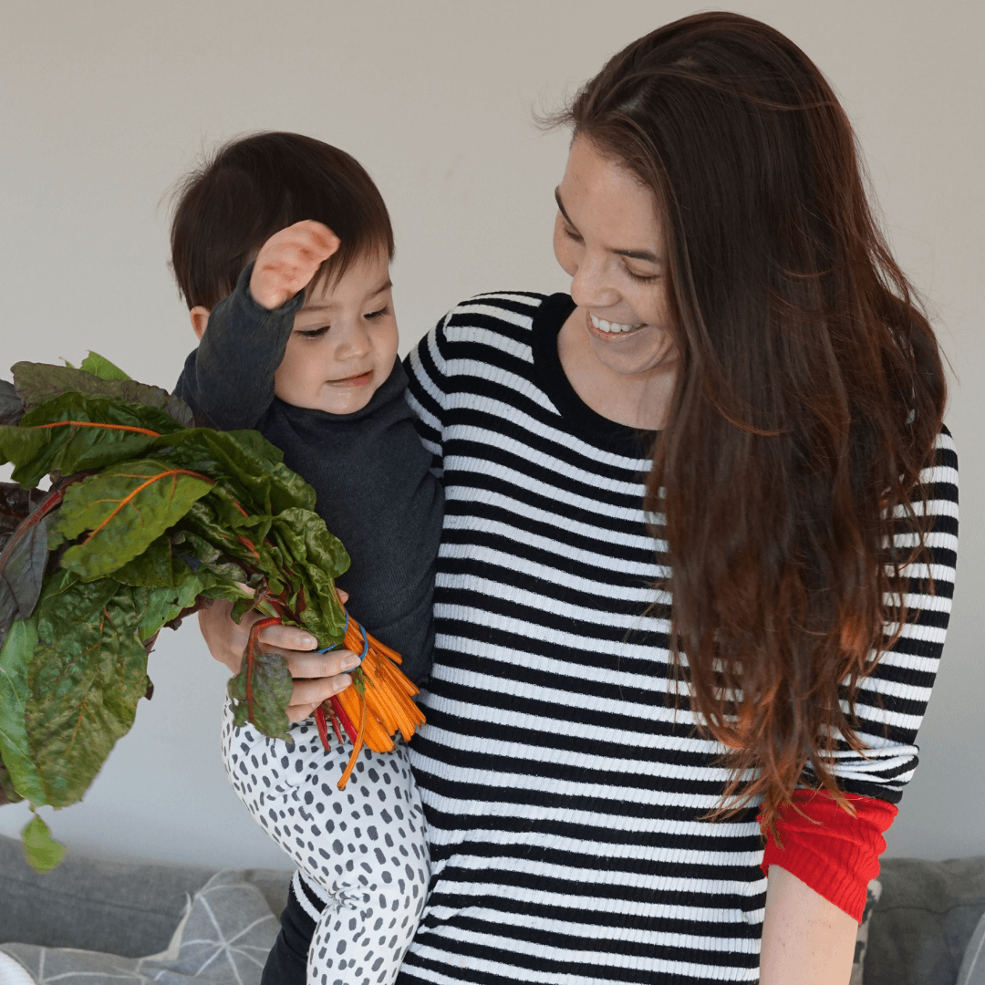 A young woman wearing a stripy top, carrying a toddler, and also holding a bunch of carrots. They are both smiling