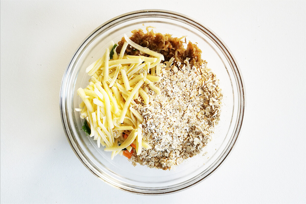 A glass bowl with grated carrot, grated aubergine and chopped spinach, grated cheese and oats