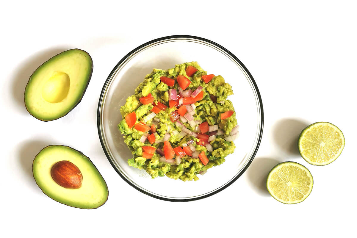 A glass bowl with smashed avocado, chopped red onion and tomatoes, next to a halved avocado and halved lime