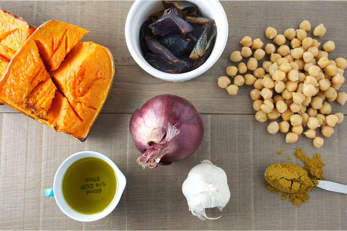 A table with half a butternut squash, red onion, fried red onion, chickpeas,, a garlic bulb and a spoon of garam masala