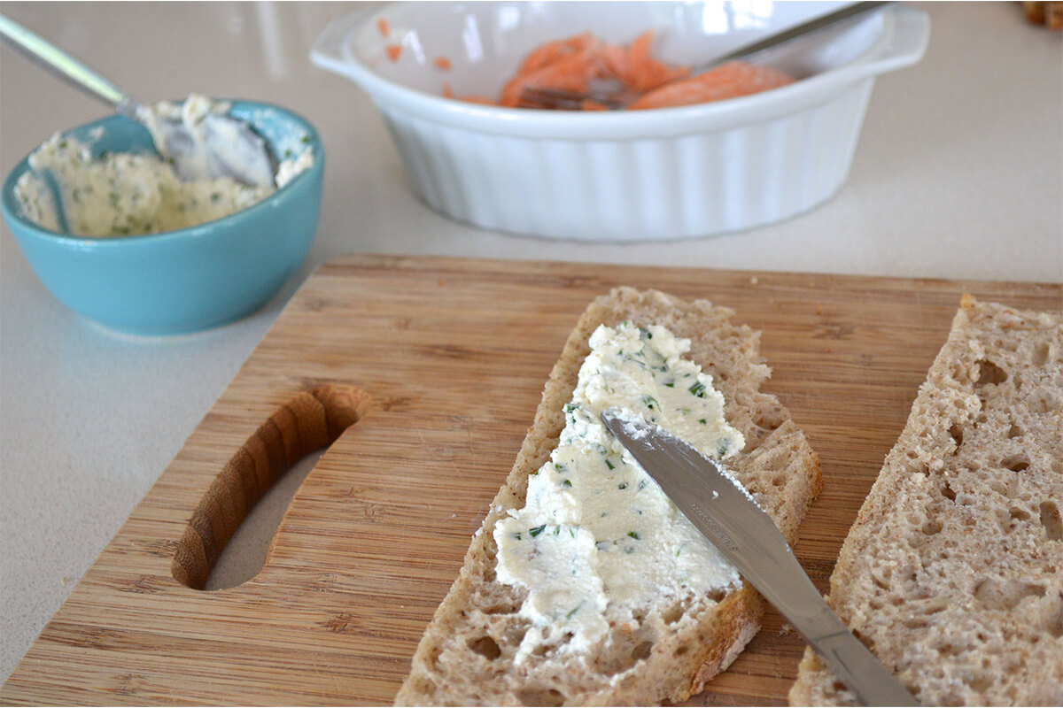 Cream cheese being spread on a slice of bread on a chopping board with a bowl of cream cheese and a dish of salmon next to it