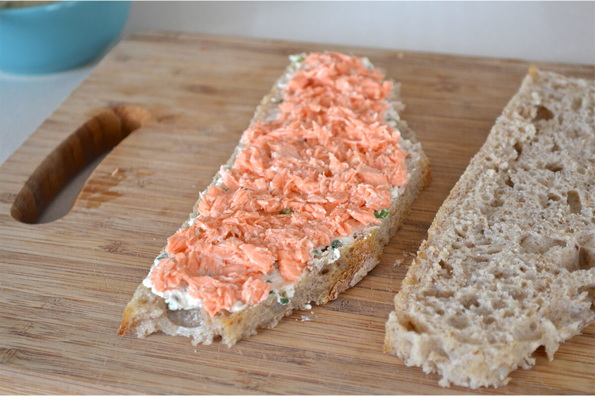 A chopping board with two slices of bread, one topped with cream cheese and poached salmon
