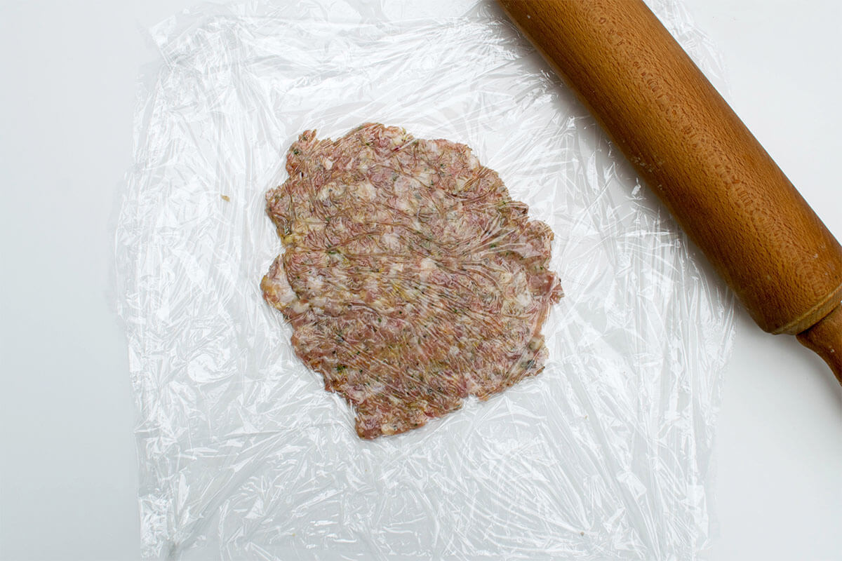 A square of cling film with meat mix rolled out onto the middle of it