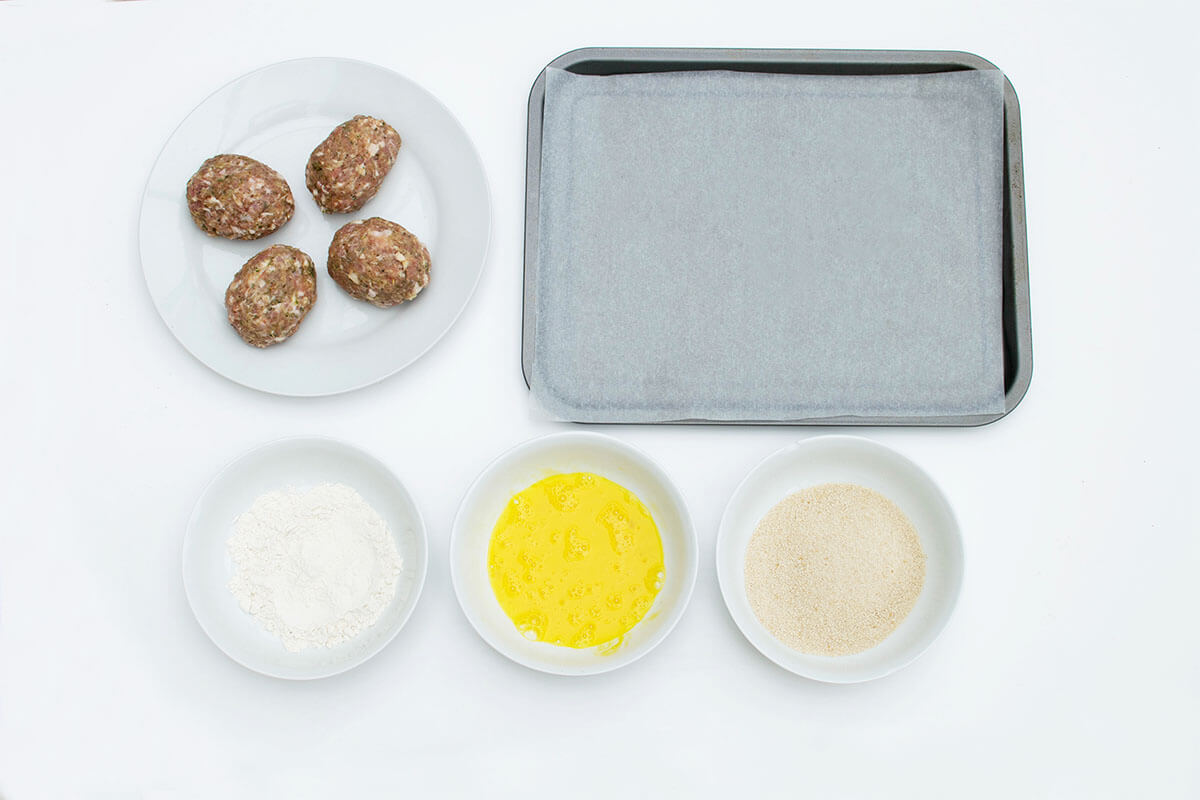 4 balls of sausage-turkey meat with eggs encased in the middle of them, next to a baking tray lined with parchment and bowls of flour, beaten eggs and breadcrumbs