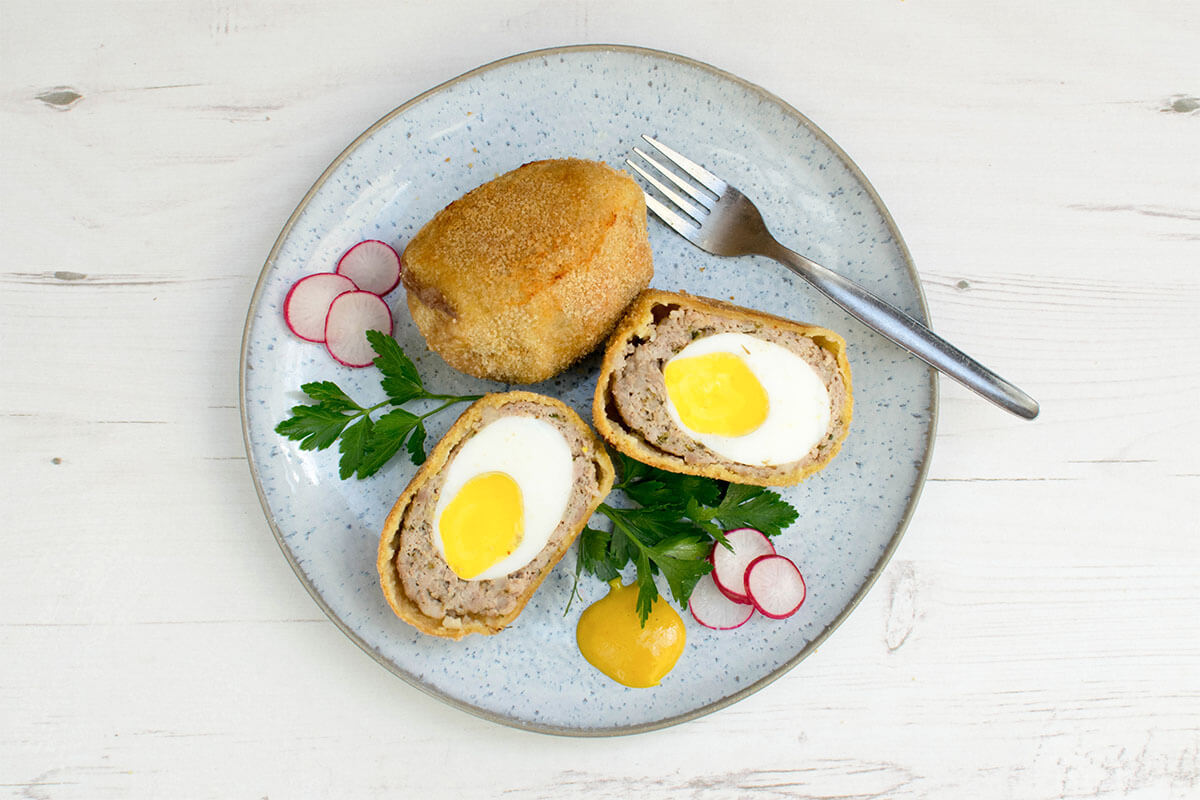 A plate with a whole and halved scotch egg, served with fresh herbs, radish and mustard