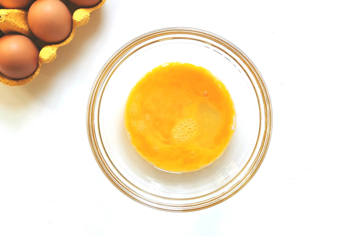 A glass bowl of whisked eggs next to a crate of eggs