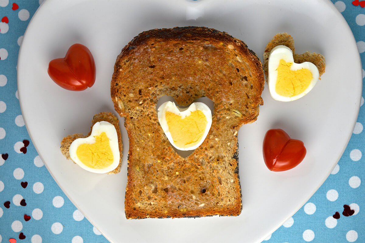 Toast with a heart shaped cut-out filled with a heart shaped boiled egg, served with 2 heart shaped boiled eggs and heart shaped tomatoes