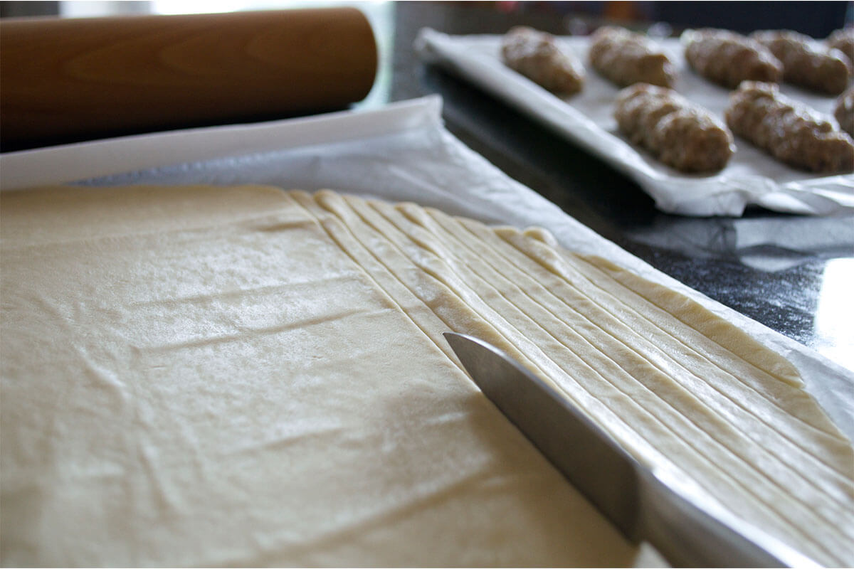 Strips of pastry being cut on baking parchment, next to a baking tray of sausages