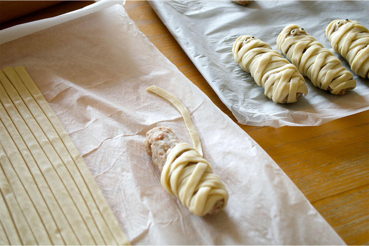 Strips of pastry wrapped around a sausage, next to a lined baking tray of sausages with strips of pastry wrapped around them