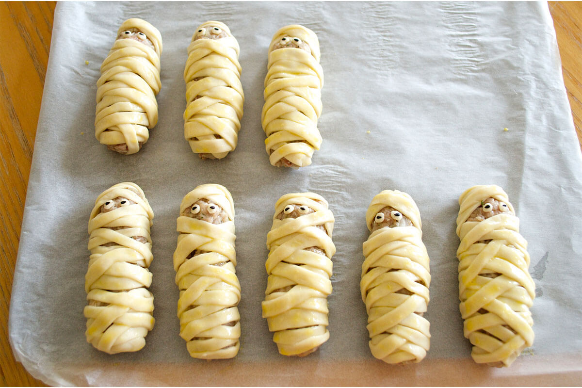 A baking tray with Mummie Sausages with sesame seed and black olive eyes