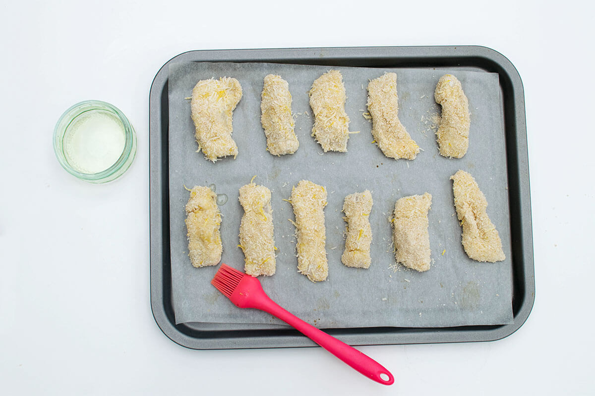 A baking tray lined with parchment paper with coated fish strips on it