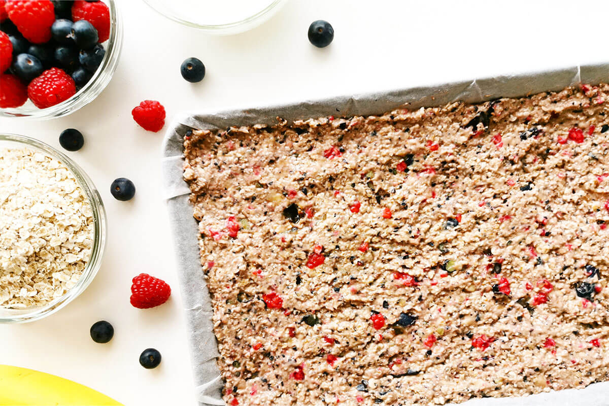 A lined baking tray with berry/banana oat mix next to a bowl of berries, a bowl of oats and a banana