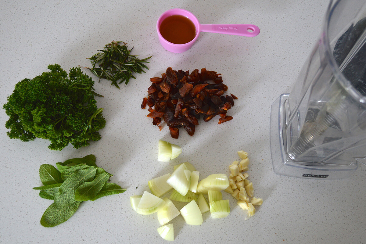 Onion, garlic, herbs and olive oil next to a food processor 