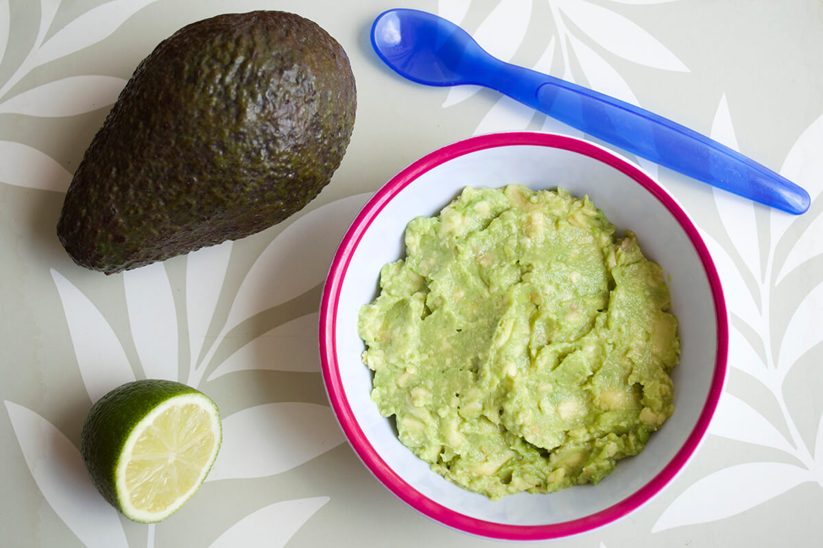 A bowl of avocado puree next to a whole avocado and half a lime