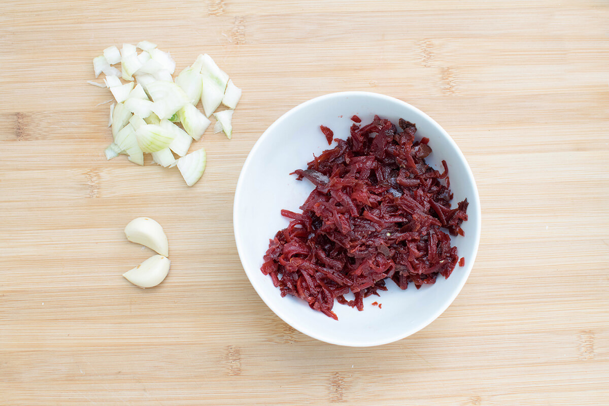A bowl of grated beetroot next to some chopped onion and 2 cloves of garlic
