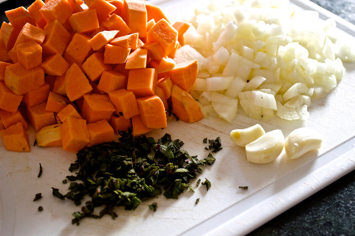 A chopping board with chopped sweet potato, onion, shredded sage leaves and 3 garlic cloves