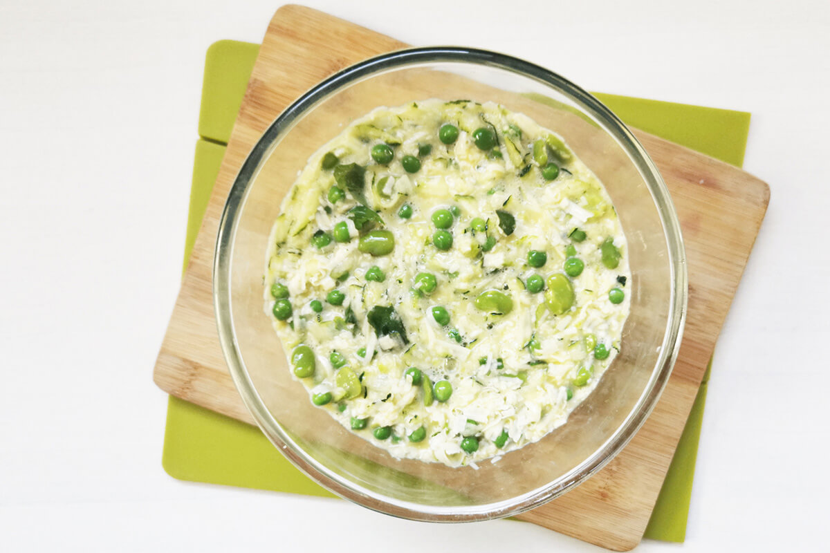 A glass bowl of eggs with grated courgette, broad beans, peas, cheese and basil