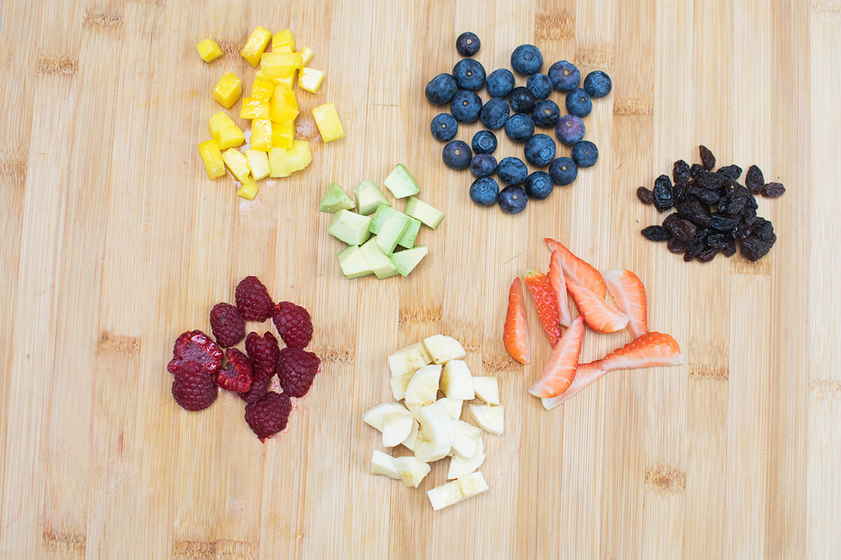 A chopping board with cut mango, avocado, strawberries, raspberries,, banana with some blueberries and raisins