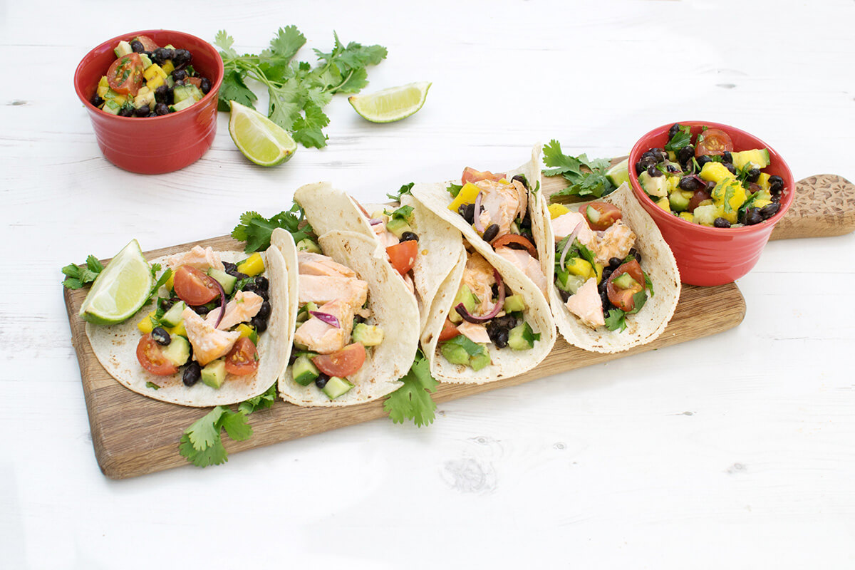 6 fish tacos on a wooden platter, served with fresh coriander, mango salsa and lime wedges