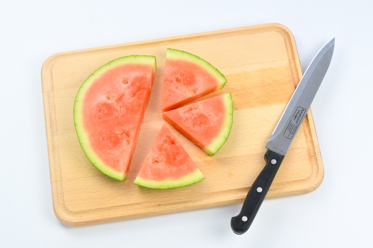 A wooden chopping board with a round of halved watermelon next to 3 large slices of watermelon