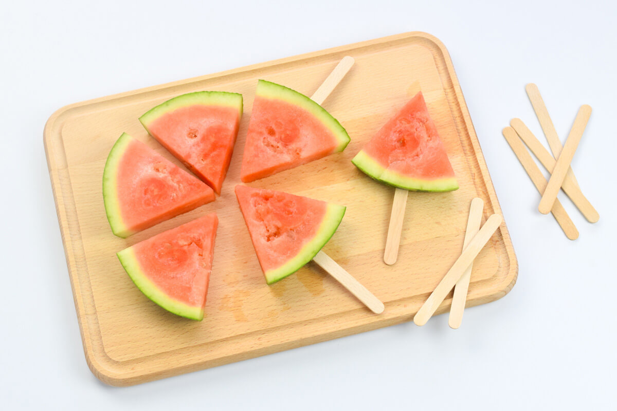 A wooden chopping board with 6 triangle shaped watermelon slices, 3 of them have wooden sticks in the rind