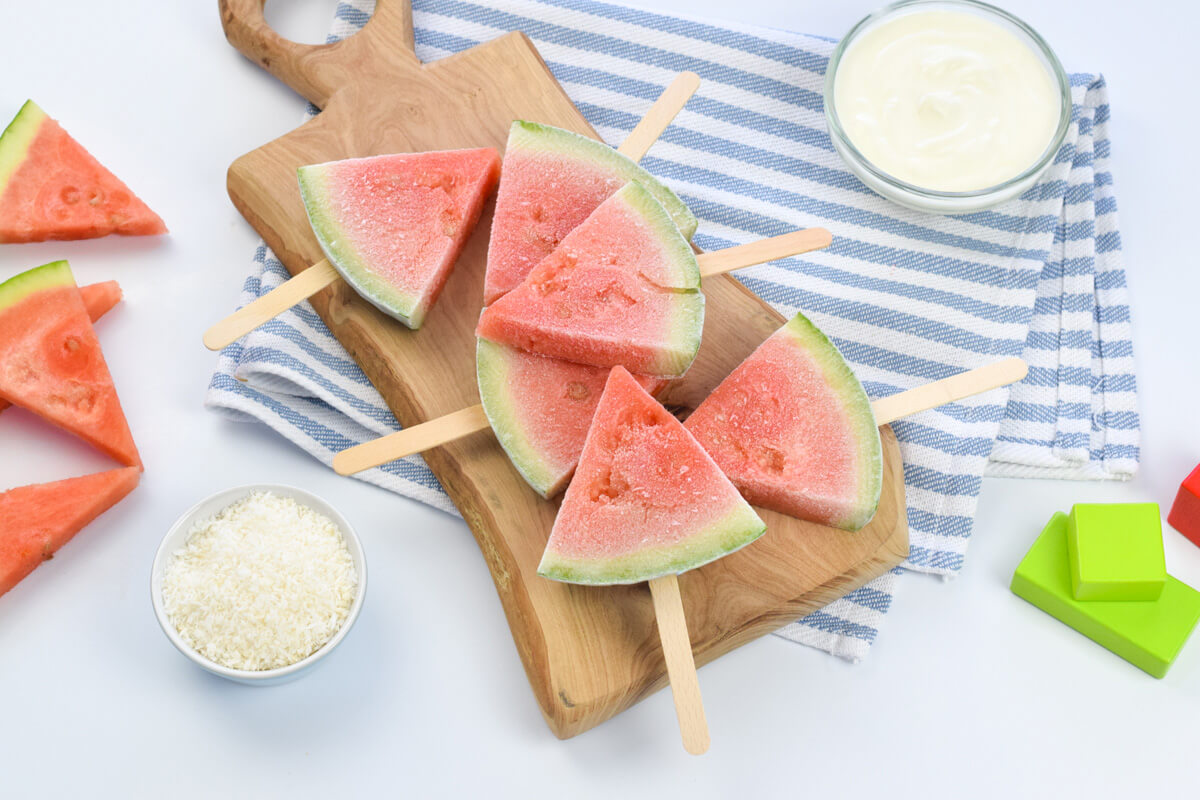 A chopping board with 6 frozen watermelon pops served with a bowl of natural yoghurt and a bowl of desiccated coconut 
