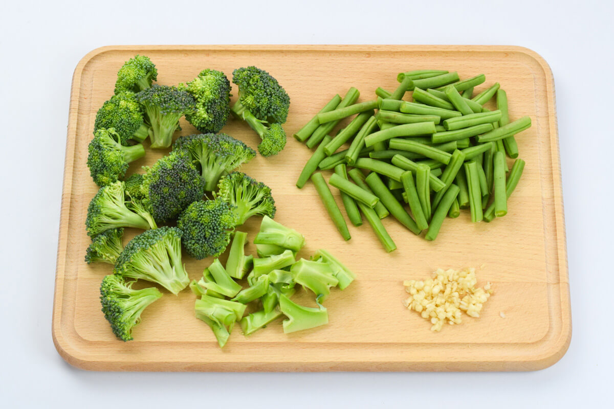 A wooden chopping board with cut broccoli florets and green beans and some peeled, crushed garlic
