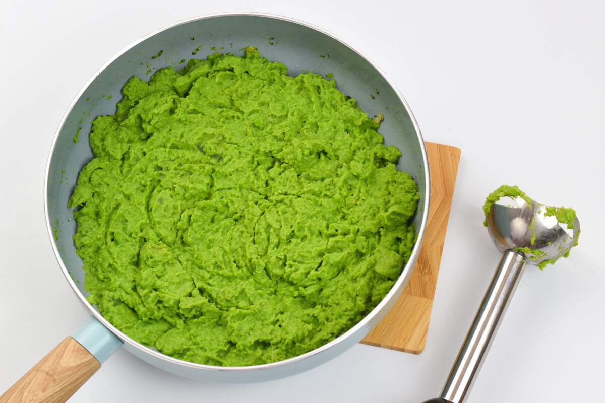 A pan of green bean, pea and broccoli mash next to a hand blender