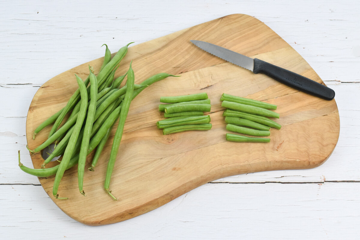 A wooden chopping board with chopped green beans next to some whole green beans