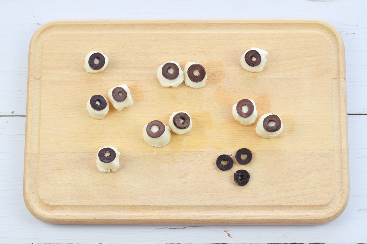 A chopping board with small mozzarella balls topped with sliced black olives to create eyes
