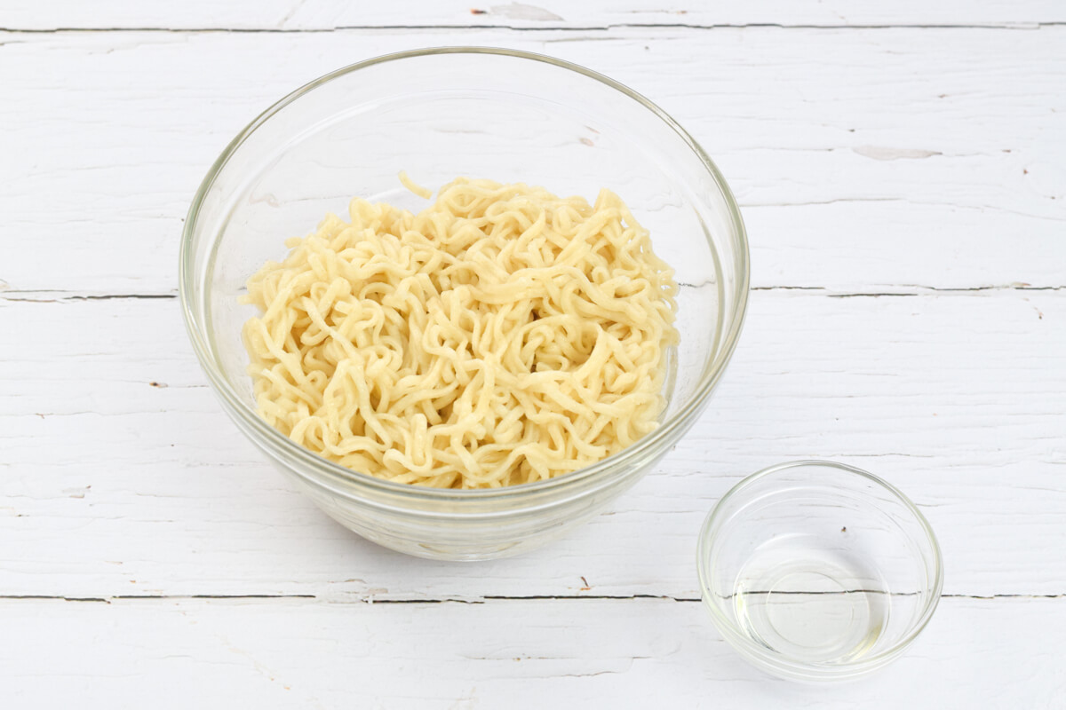 A glass bowl of noodles next to a small glass bowl of vegetable oil