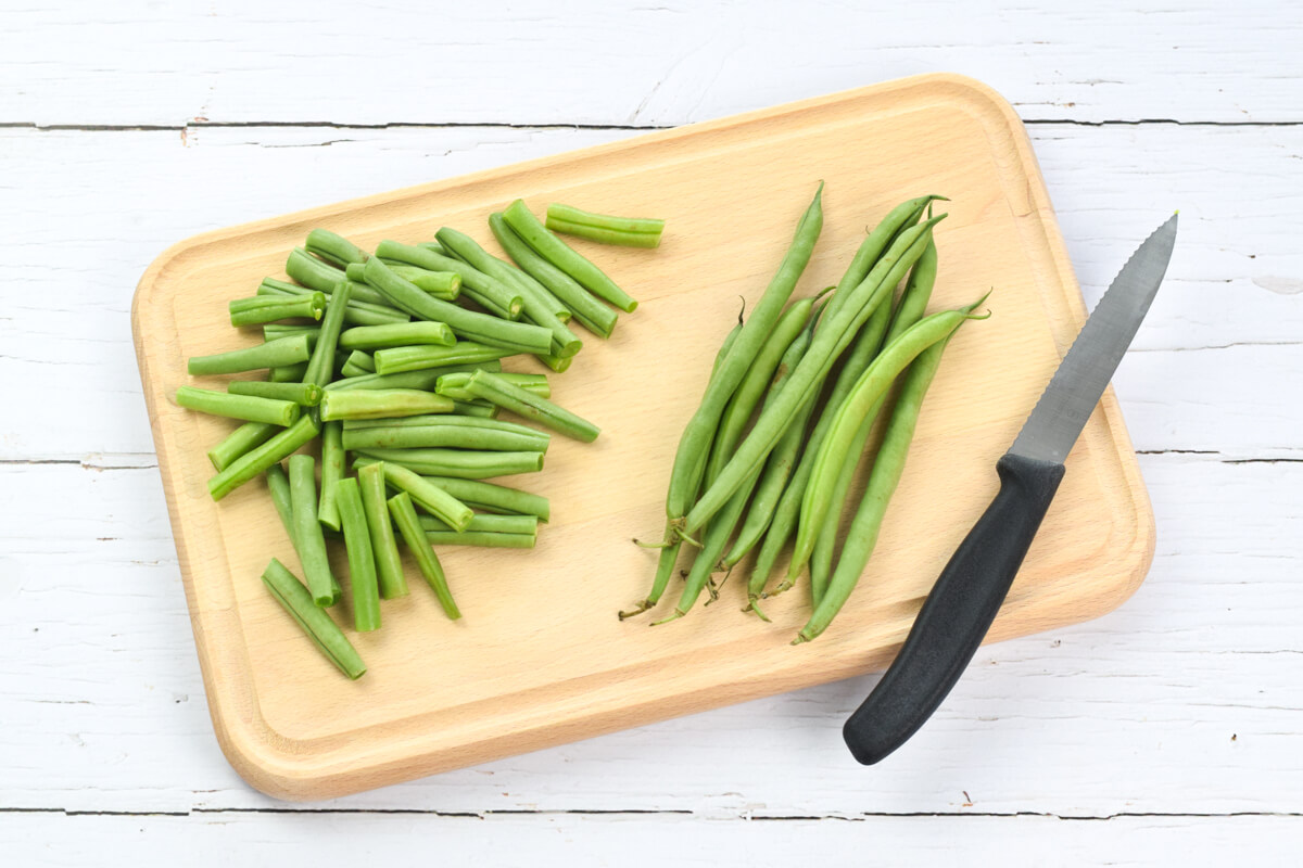A wooden chopping board with some chopped green beans, next to some whole green beans
