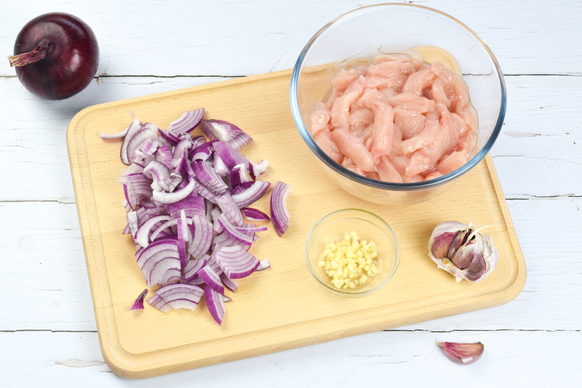 A chopping board with some chopped red onion, a glass bowl with raw chicken, a small glass bowl with garlic and some garlic cloves
