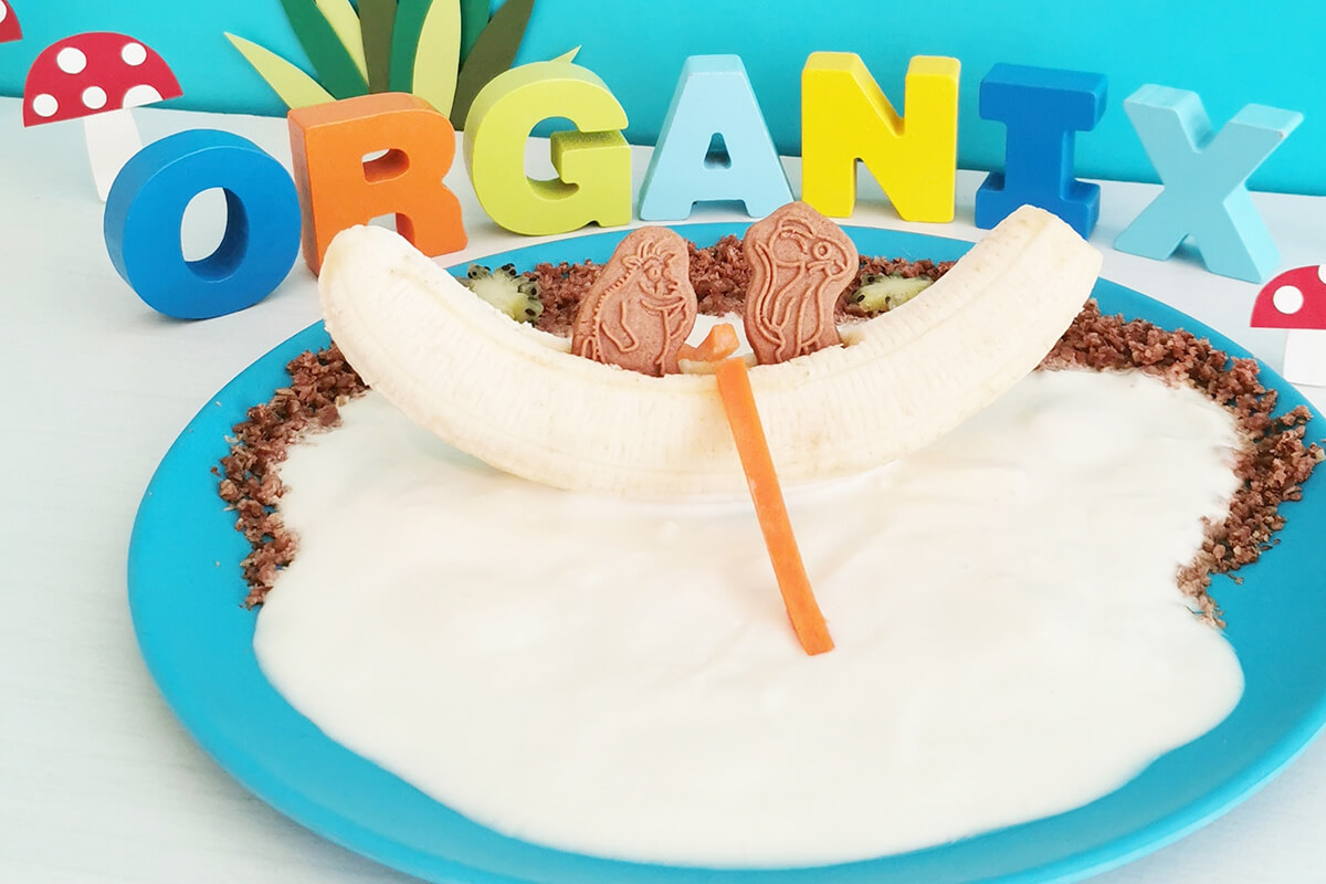 Yoghurt spread on plate with Oaty Bar crumbled and sprinkled around the edge and a banana on top, creating a boat and 2 Grufallo biscuits in the banana creating boat passengers. Sliced carrot sticks on either side create oars
