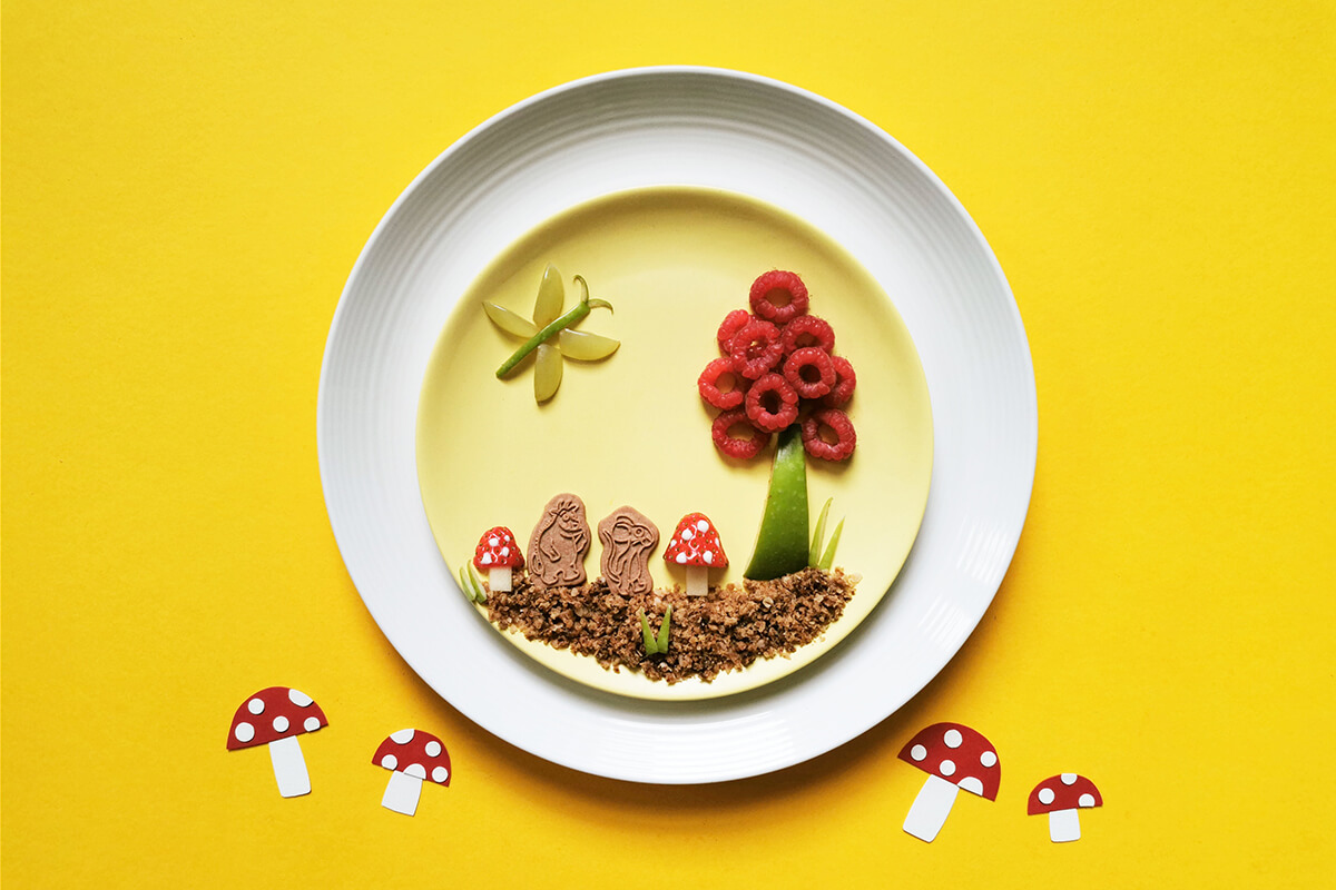 Gruffalo Biscuit Fun Plate, decorated with crumbled biscuits, an apple slice and raspberries to make a tree,  apple chunks and strawberries to make 2 toadstools, with 2 Gruffalo biscuits in between the toadstools  and an apple slice and grapes pieces to make a butterfly 